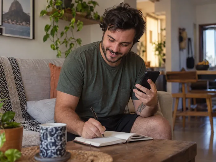 Homem no sofá anotando num caderno com o celular na mão
