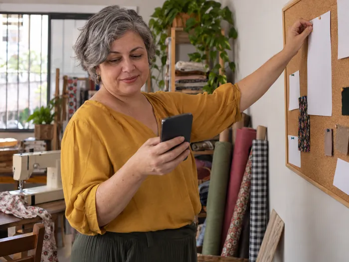 Mulher de blusa mostarda no ateliê de costura de casa, prendendo um papel num quadro de cortiça com o celular na outra mão